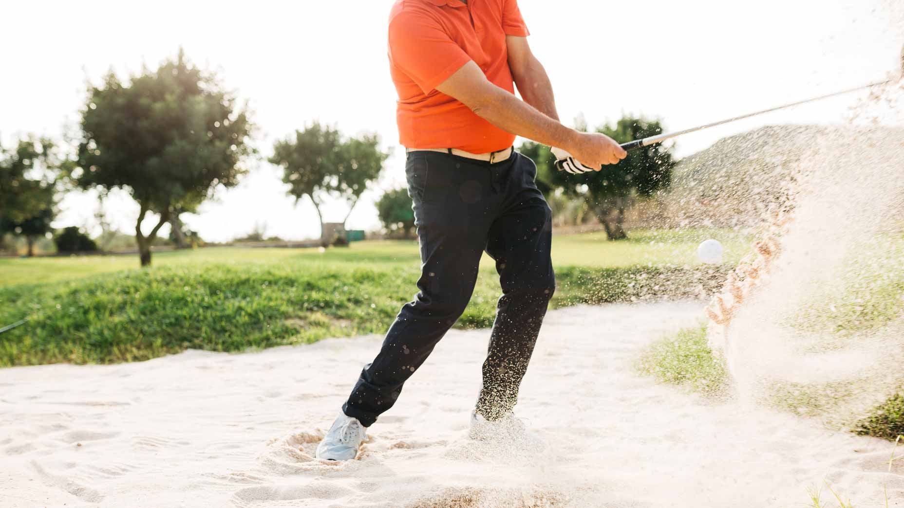 Golfer in orange shirt hits a bunker shot on a golf course.